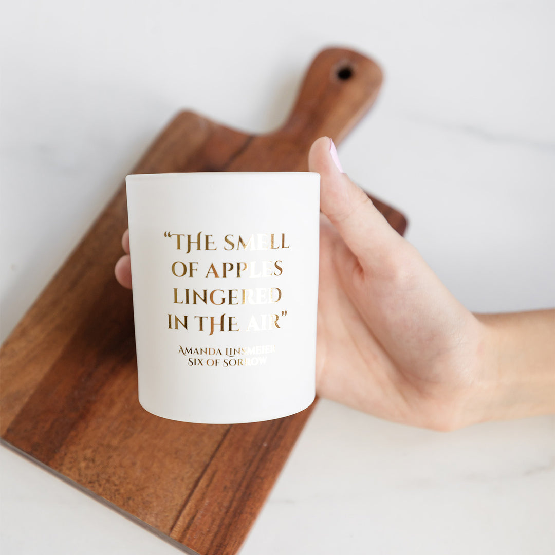 A hand holds a white candle above a wooden cutting board. On the candle is the following text in gold lettering: The smell of apples lingered in the air. Amanda Linsmeier Six of Sorrow
