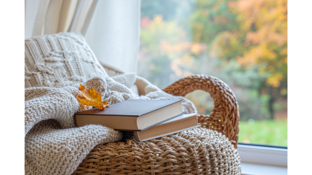 Cozy wicker chair by a window with two stacked books and a knitted cream blanket, autumn leaves visible outside.