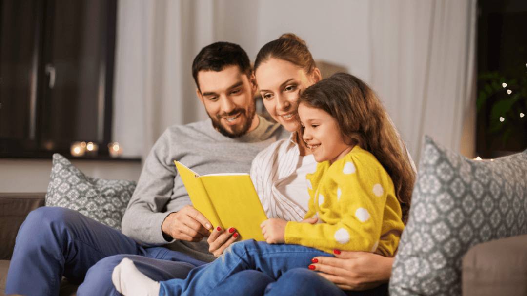 A happy family sitting on a couch reading a yellow book together in a cozy living room. Illustration of the words "reading goals" in playful pink and peach fonts on a pale background.