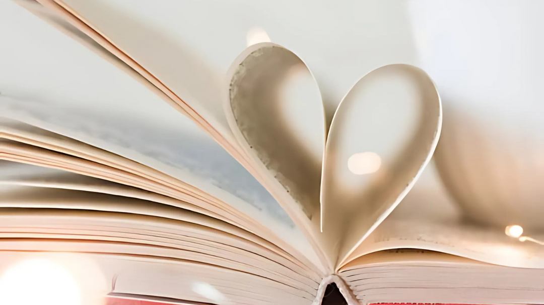 An open book with pages folded into a heart shape, illuminated by soft fairy lights, sits on a cozy surface with a blurred cup in the background.