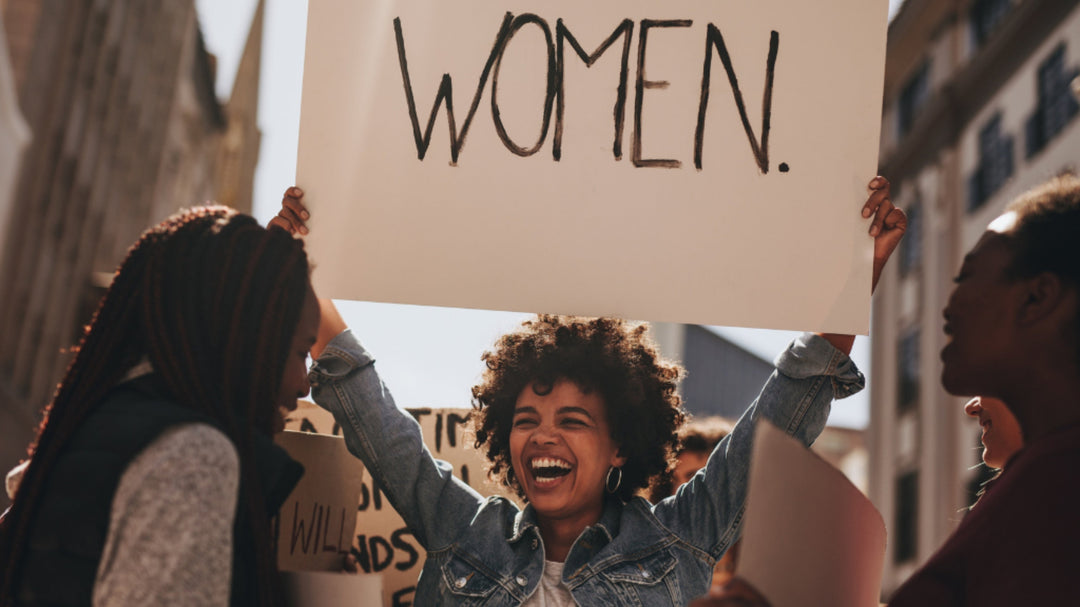 Smiling woman at a protest holding a large sign that says “WOMEN” while surrounded by others in an urban setting.