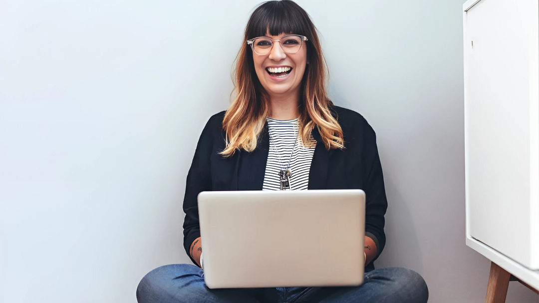 A woman sitting on the floor with her laptop, smiling and wearing glasses, conveying a positive and engaging online experience.