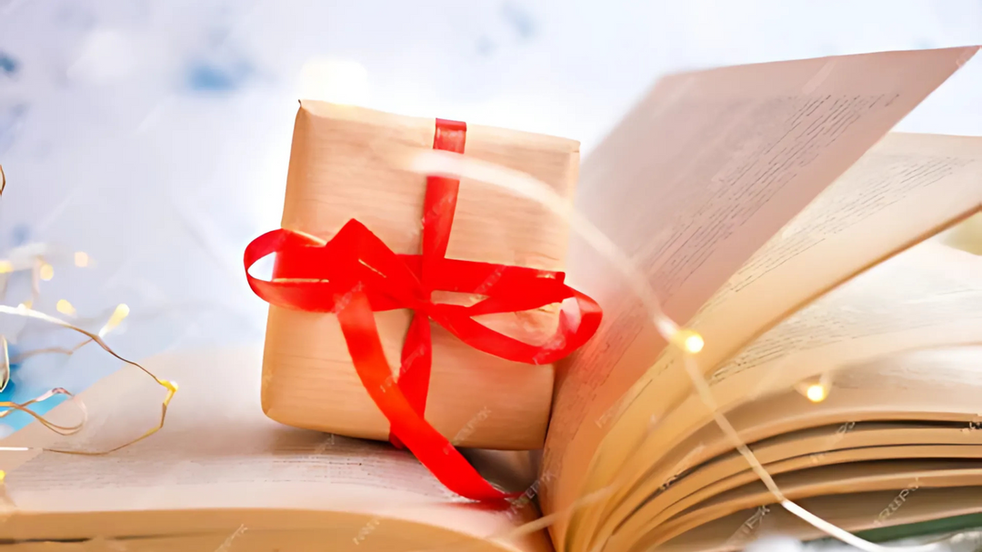 An open book with a small gift wrapped in brown paper and tied with a red ribbon placed on its pages, surrounded by fairy lights.