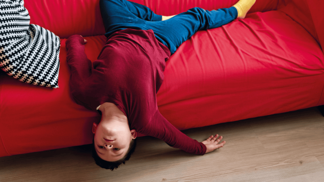 Boy lying upside down on a bright red couch, looking bored with his head hanging toward the floor.