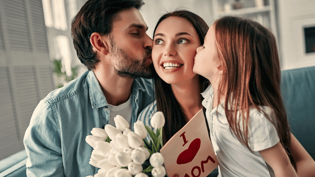 A happy mother sits on a couch holding white tulips and a handmade "I ❤️ Mom" card, kissed on each cheek by her husband and young daughter.