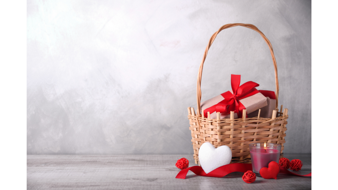 Wicker gift basket with a wrapped present tied in red ribbon, candle, and heart decorations styled for Valentine’s Day.