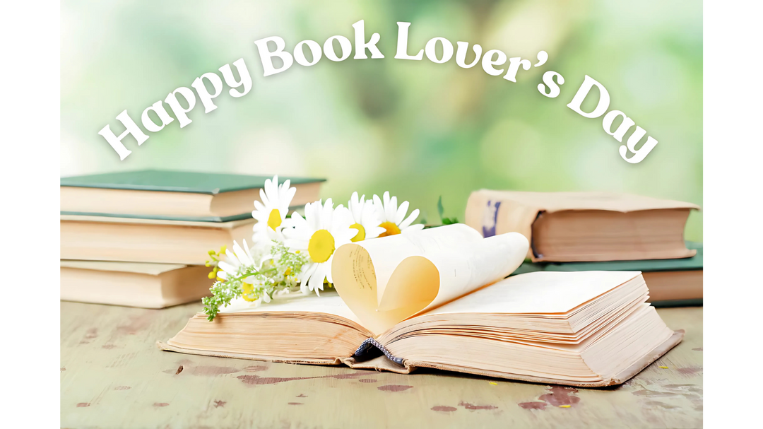 Open book with heart-shaped pages, daisies, and stacked books on a wooden table. Text reads 'Happy Book Lover's Day.