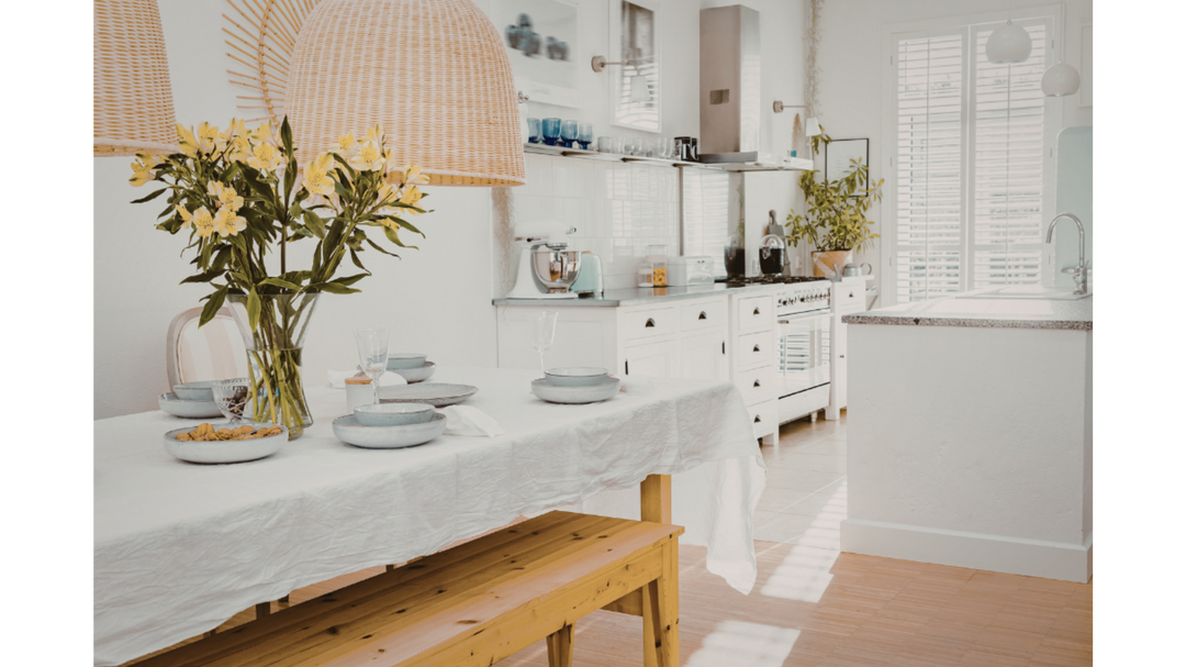  Bright, airy white kitchen and dining area with a wooden table set with bowls, flowers in a vase, and woven pendant lighting.