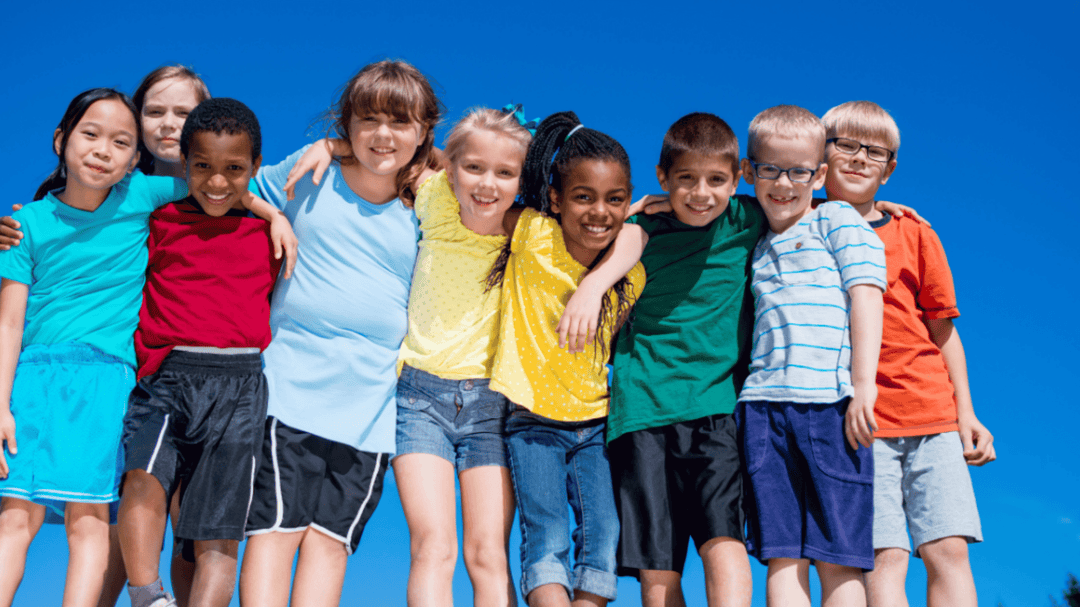 Group of smiling children standing in a row outdoors, arms around each other, against a bright blue sky.