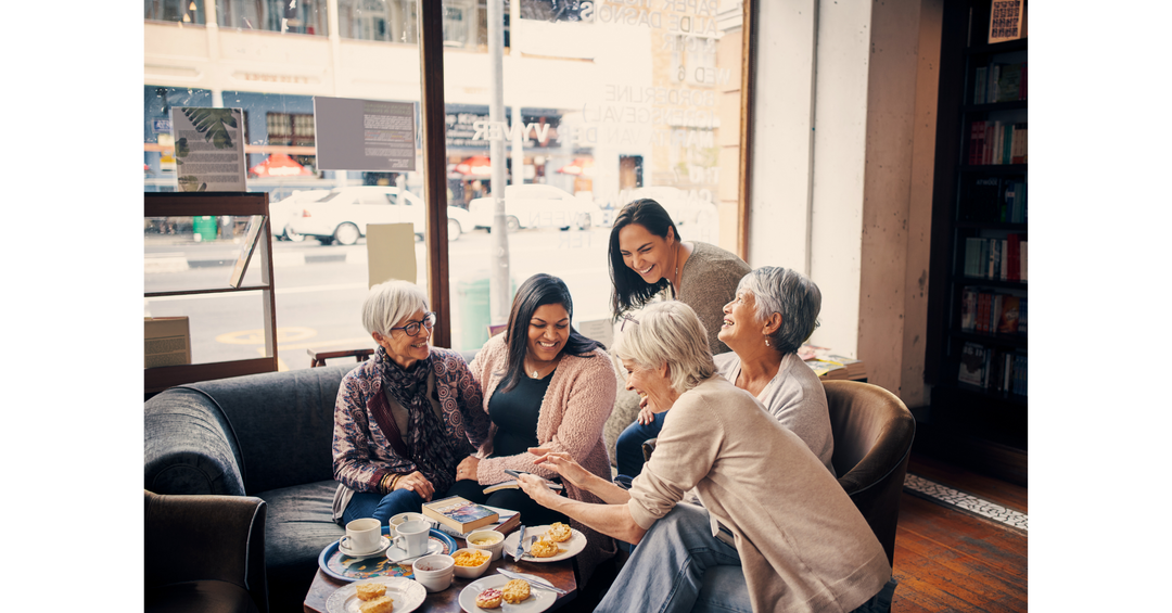 Group of women laughing and chatting together in a cozy café setting with books, tea, and snacks on the table.