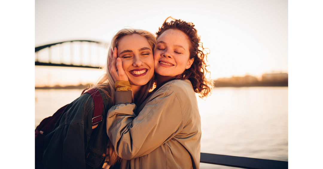 Two smiling friends hugging by the water at sunset, creating a warm and heartfelt summer moment.