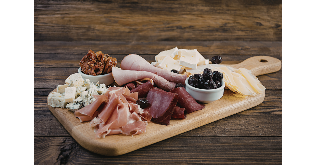 A wooden charcuterie board with cured meats, cheeses, olives, and sun-dried tomatoes on a rustic wooden table.