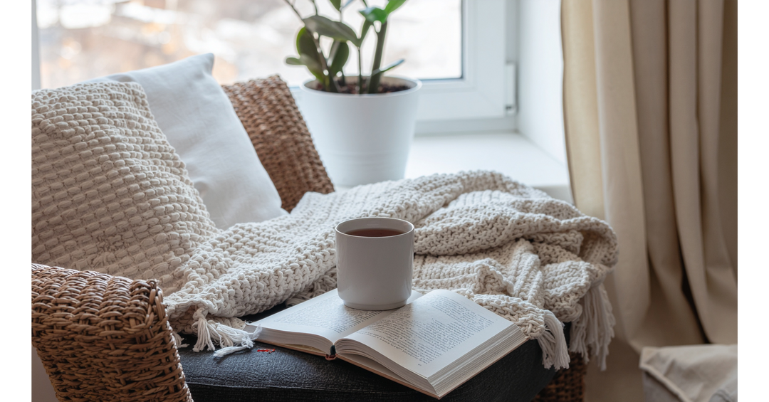 Cozy reading nook with a knitted blanket, open book and warm drink beside a bright window.