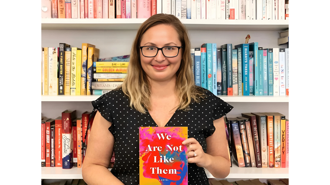 A woman holding the book "We Are Not Like Them" in front of colorful bookshelves, smiling and wearing a black polka dot top.