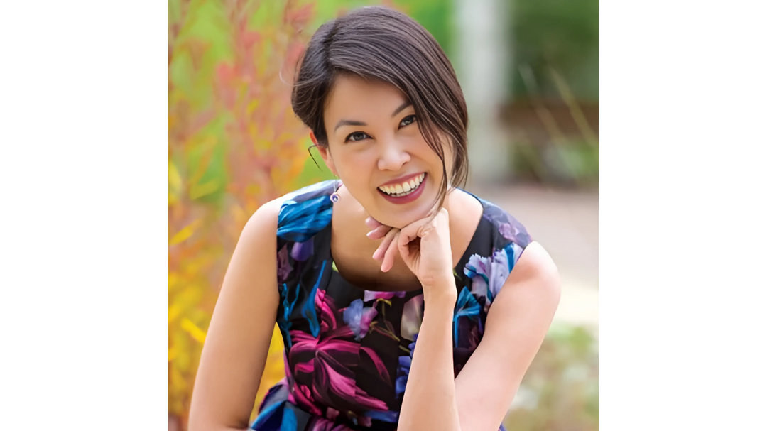 Stacey Lee, smiling and resting her chin on her hand, wearing a colorful dress, with a blurred outdoor background.