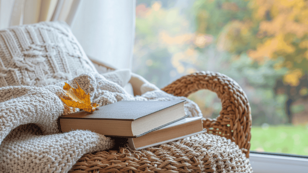 Cozy wicker chair with a knit blanket, two books, and an autumn leaf by a bright window.