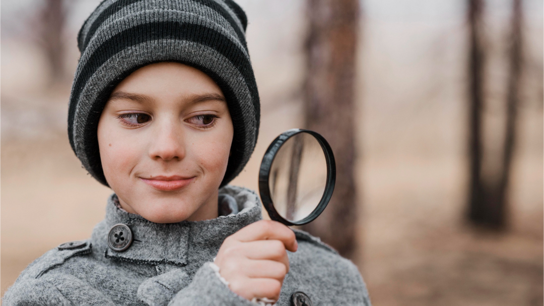 A young boy in a winter coat and beanie hat, holding a magnifying glass and smiling while exploring outdoors.