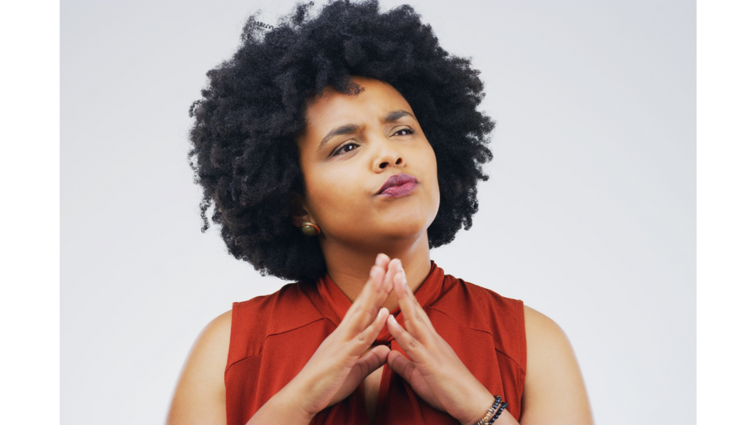 Woman with natural curly hair in a red sleeveless top, looking thoughtful with hands clasped under her chin against a neutral background.