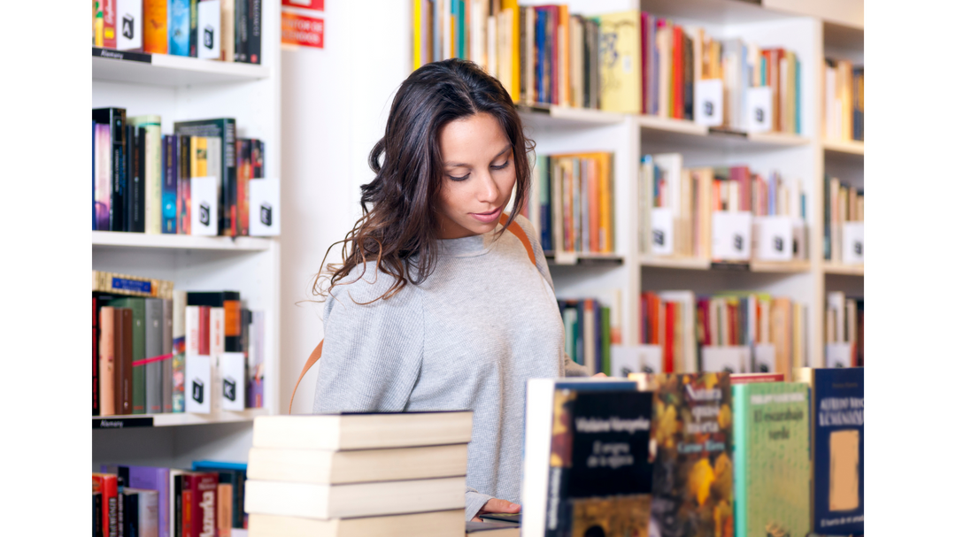 A woman with long brown hair wearing a gray sweater is browsing books in a bookstore, surrounded by shelves filled with colorful books and neatly stacked books on a table in front of her.