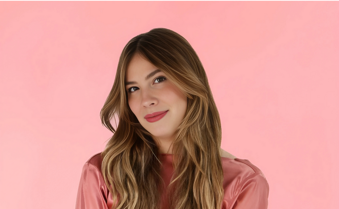 Emily Henry, author, posing against a soft pink background wearing a silky pink top, her hair styled in loose waves, smiling warmly.