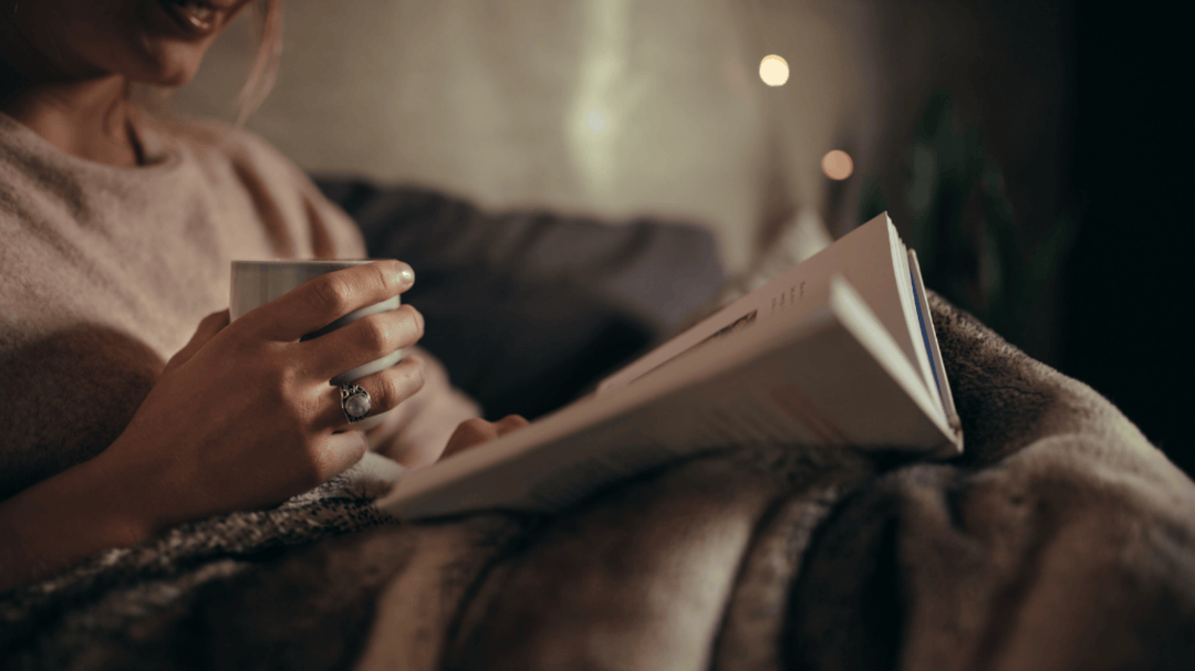 Woman snuggled under a blanket reading a book and holding a warm drink, cozy atmosphere.