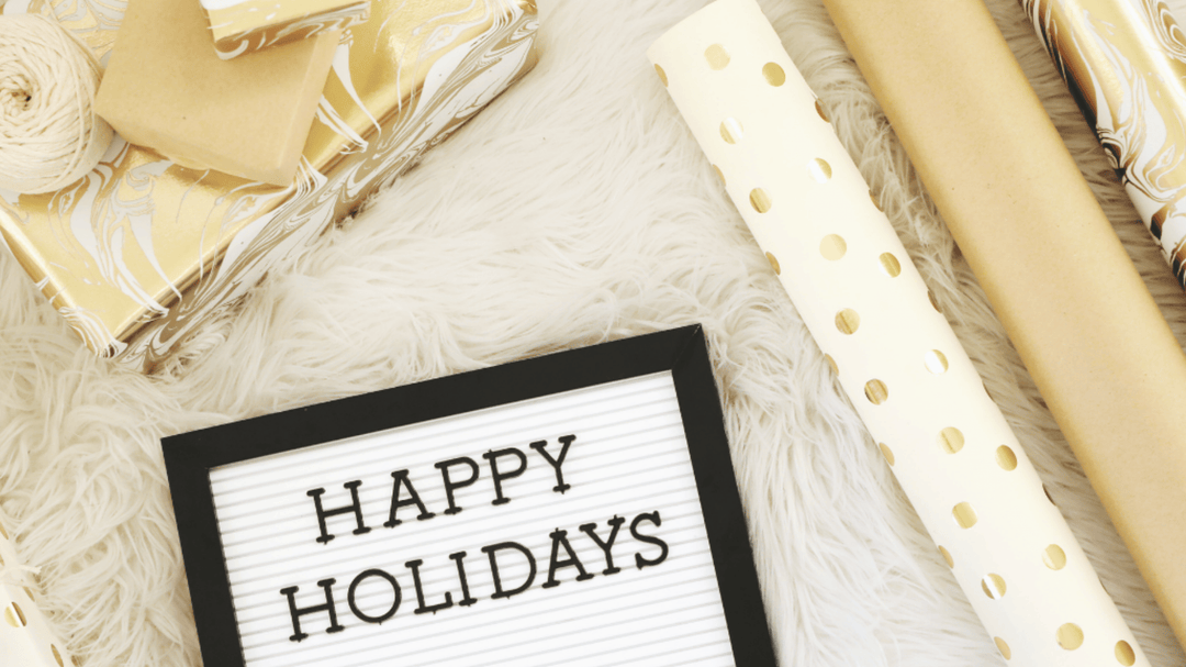 Festive flatlay with gold-wrapped gifts, wrapping paper, and a letterboard reading 'Happy Holidays'.