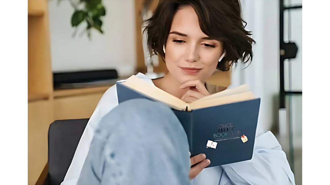 A woman with short dark hair, wearing a light blue shirt, sits comfortably while reading a book with a focused expression in a cozy room.