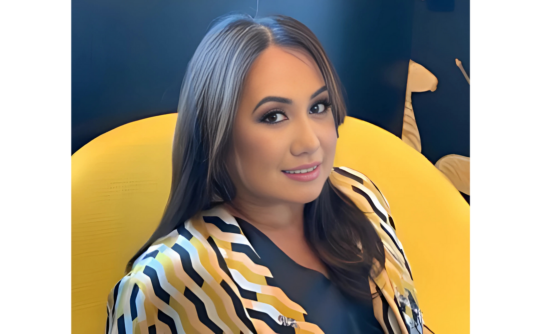 Melissa de la Cruz, a renowned author, smiles while seated in a bright yellow chair, dressed in a stylish striped outfit against a navy background.