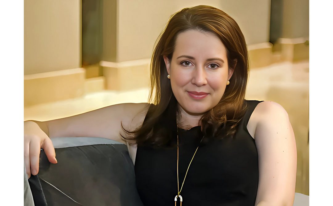 Author Julia Quinn with shoulder-length brown hair, wearing a black sleeveless top and a gold necklace, is sitting comfortably on a sofa, smiling.