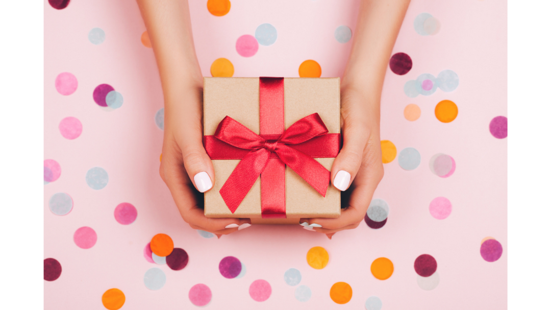 Hands holding a small kraft gift box wrapped with a bright red ribbon against a pink background with colorful confetti dots.