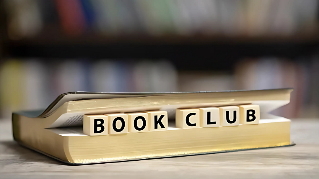 An open book on a table with letter tiles spelling "Book Club" placed across the pages, with a blurred bookshelf in the background.