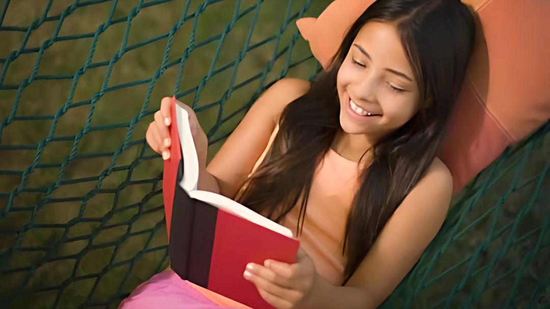 A smiling girl relaxes on a hammock outdoors, reading a red book and enjoying the serene, sunny atmosphere.