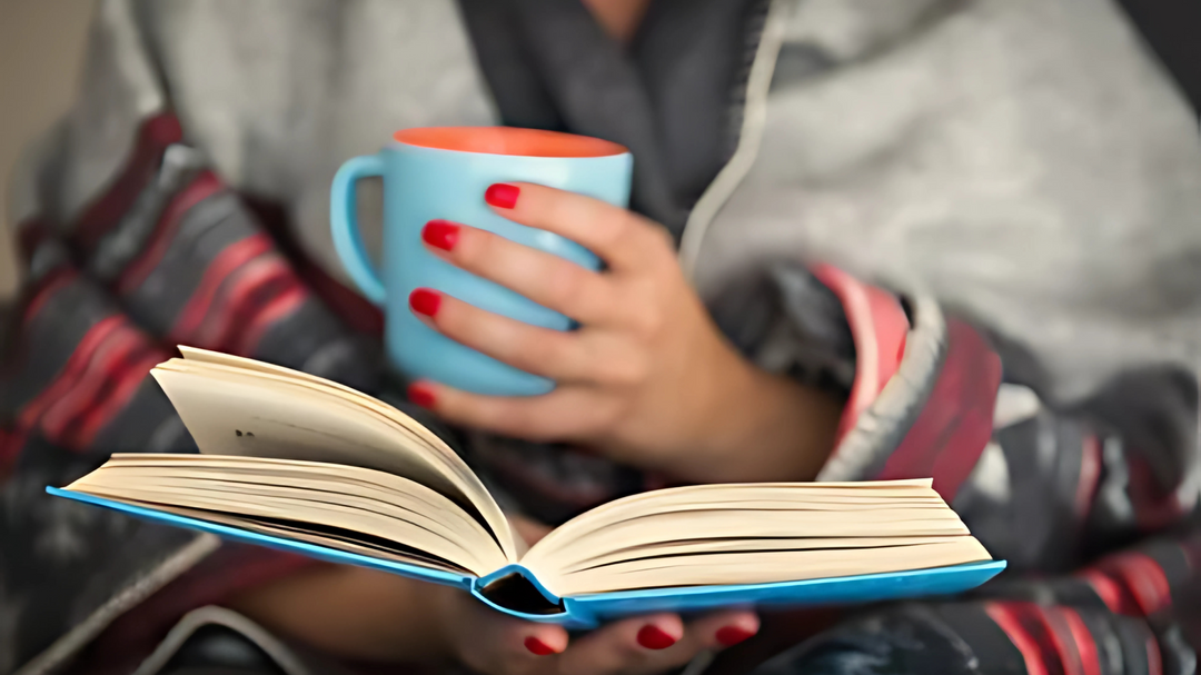 A person wrapped in a cozy patterned blanket holding a blue mug and reading an open book, with red-painted nails in focus.