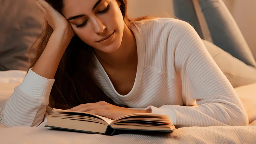 A woman relaxing on a bed in soft lighting, engrossed in reading a book, wearing a comfortable white long-sleeve top and jeans.