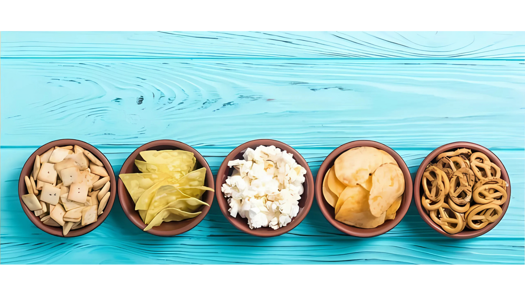 Five bowls of assorted snacks including crackers, chips, popcorn, and pretzels arranged on a vibrant blue wooden surface.