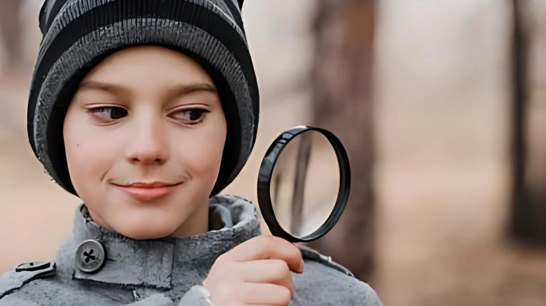 A young boy wearing a gray coat and striped beanie holds a magnifying glass, smiling as he examines something outdoors, evoking curiosity.