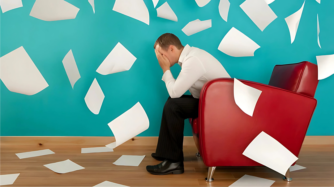 A man sits on a red chair with his face in his hands while white papers float around a turquoise background, suggesting stress.