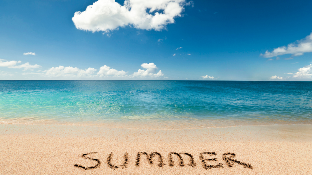 The word “SUMMER” is written in the sand as turquoise waves lap at the shore beneath a bright blue sky and fluffy clouds.
