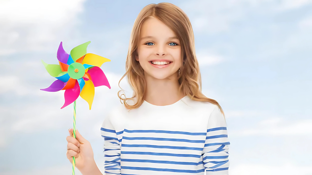 Smiling girl holding a colorful pinwheel, wearing a white and blue striped shirt, with a blue sky background