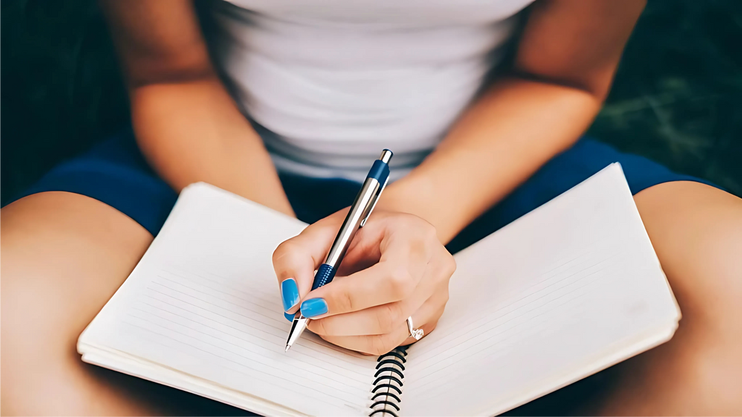 A close-up of a person writing in a spiral notebook with a blue pen, wearing a white top and blue nail polish, seated outdoors.