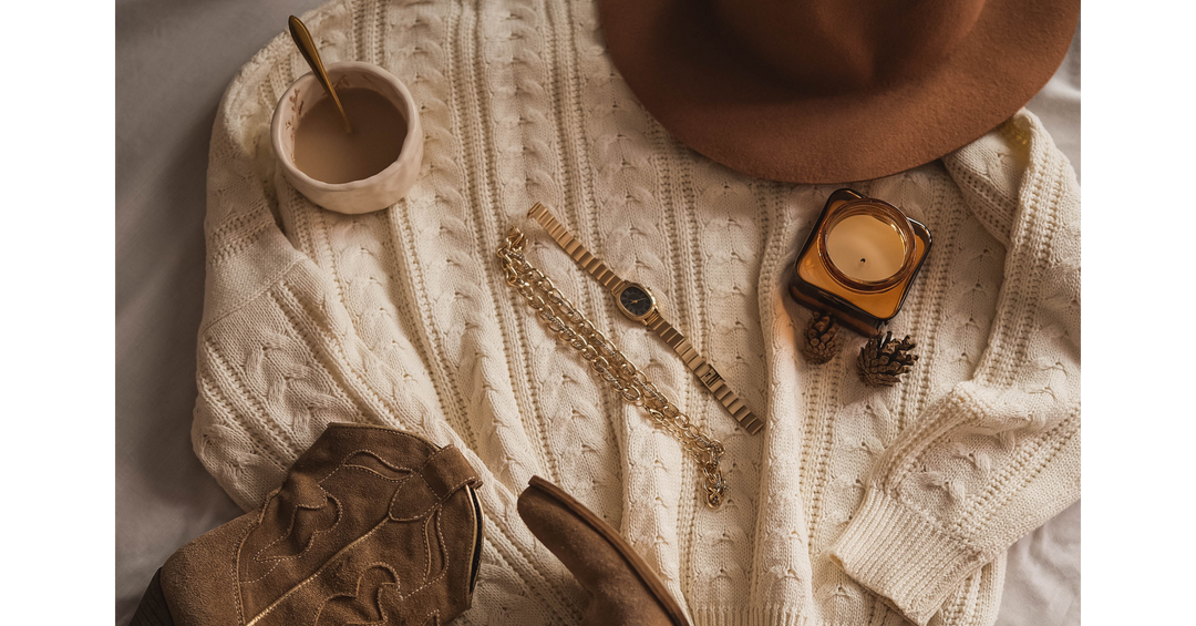 Cream cable-knit sweater styled with a brown hat, suede boots, gold watch, layered necklace, coffee, and candle for a cozy autumn look.