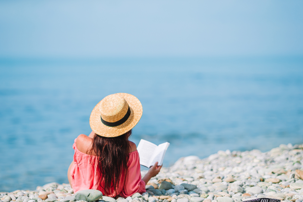 A young woman wearing a straw hat and pink off-shoulder top sits on a rocky beach, reading a book while facing the calm blue sea.