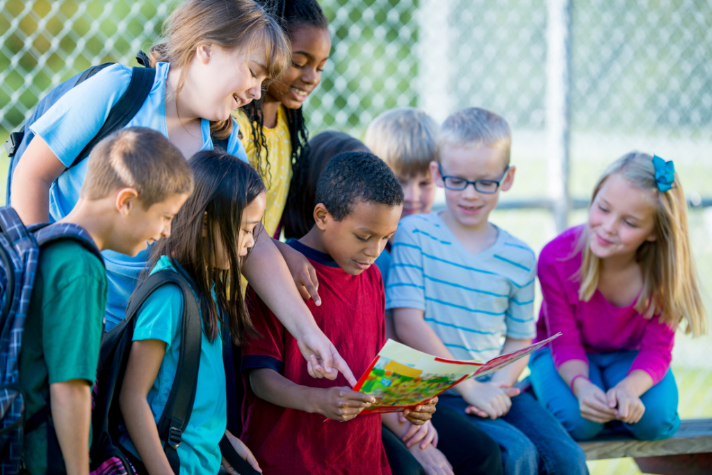 A diverse group of school children gathered outside, smiling and reading a colorful book together near a playground fence.