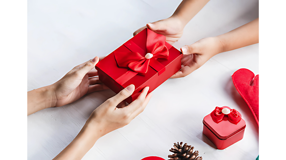 Two hands exchanging a red gift box with a bow, surrounded by holiday decorations including pinecones and a smaller red gift box.
