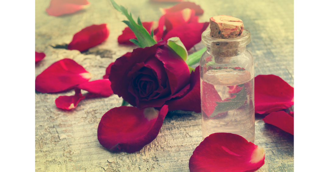 A small glass bottle of rose-scented perfume oil resting amongst scattered red rose petals and a fresh red rose, creating a romantic, floral atmosphere perfect for a romantasy-inspired reading experience.