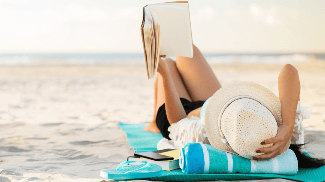 Woman reading a book on the beach with a hat, towel, and flip-flops beside her on a turquoise mat.