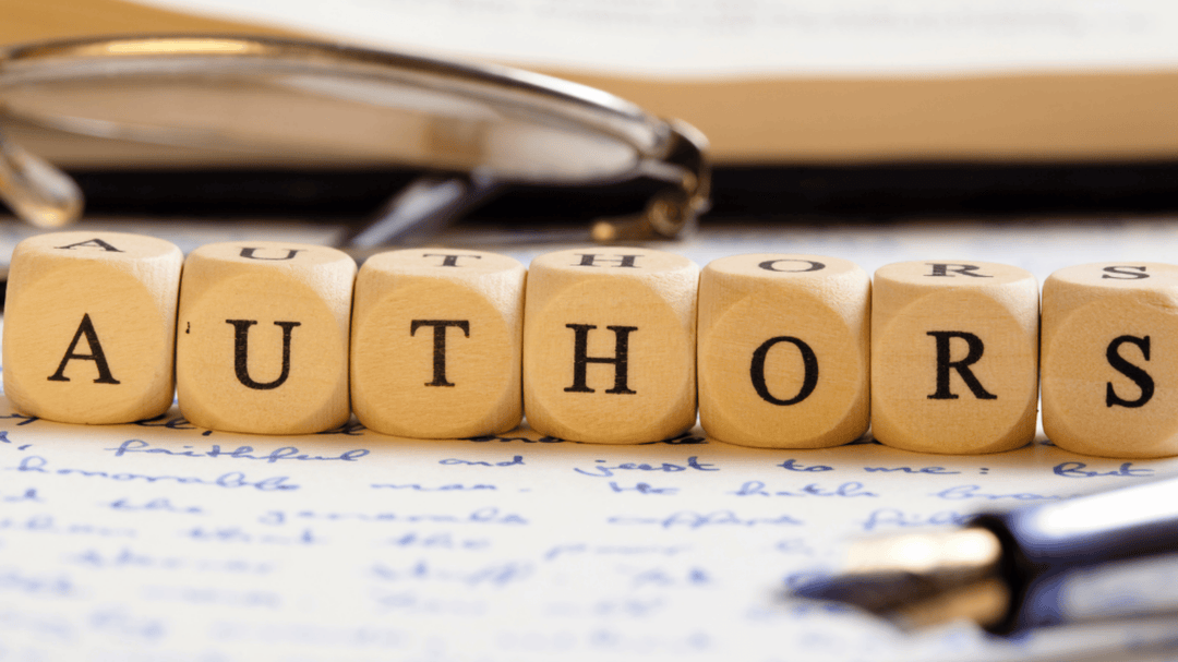 Wooden letter blocks spelling “AUTHORS” arranged on a handwritten page with a pen and reading glasses in the background.