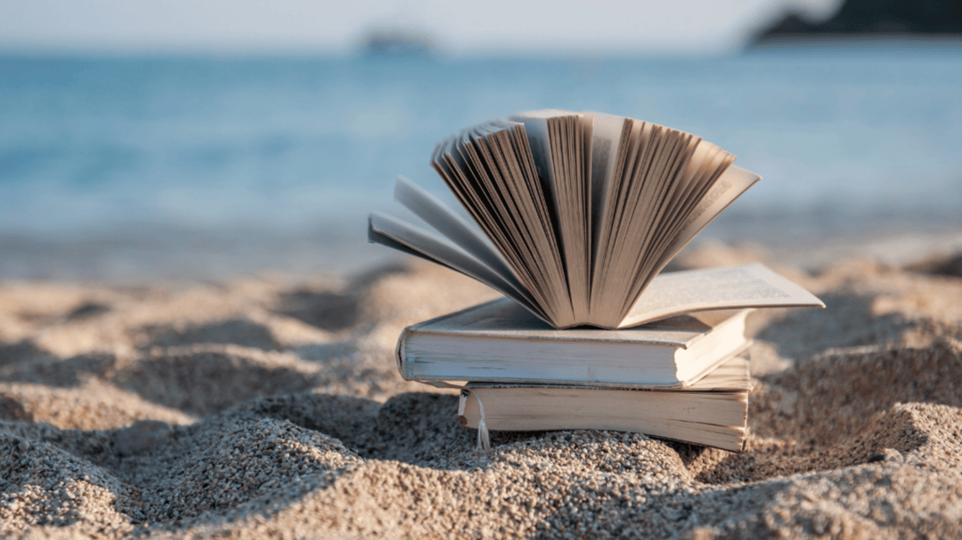 Stack of books on a sandy beach with an open book fluttering in the breeze near the ocean.