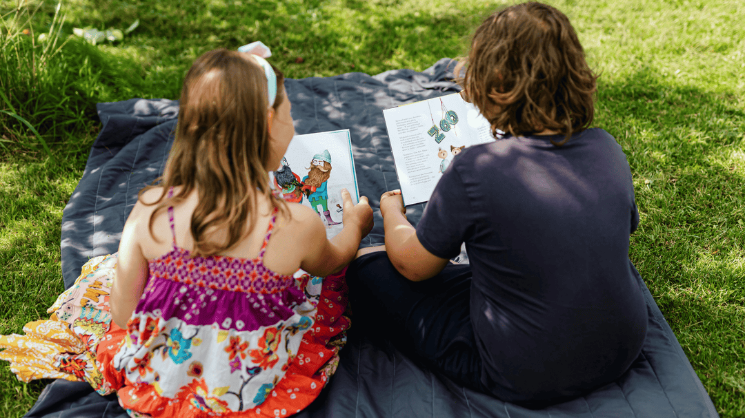Two kids sit on a blanket outside, reading colorful books together on a sunny day.