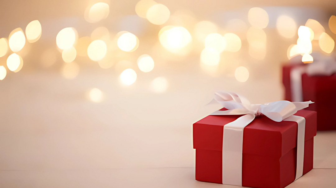 A red gift box with a white ribbon sits on a softly lit surface, surrounded by a warm background of golden holiday lights.